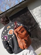 Mochila de cuero negro, con forro interior de lino y herrajes de bronce. Presenta un bolsillo frontal con cierre y un parche de cuero marrón con logo grabado.