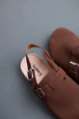 Sandalias marrones estilo zueco de hombre, con tira ajustable en el empeine y en el talón con hebillas metálicas. Presentan suela de corcho y base de goma estriada.