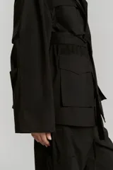 Parka negra de gabardina con corte amplio, cuello camisero, manga amplia con recortes y presillas en puños, cartera de botones oculta, bolsillos plaque con tapa, canesú con pestaña en delantero y cinturón con medialuna metálica.
