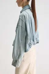 Sobrecamisa oversize de denim celeste con acabado lavado, cuello clásico, cierre frontal con broches metálicos, bolsillos frontales con solapa, mangas largas con puños abotonados y detalle elastizado en el ruedo posterior.