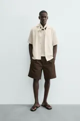 Camisa de calce relajado confeccionada en tejido con algodón de color beige. Presenta cuello bowling, manga corta, aberturas laterales en el bajo y cierre frontal con botones.