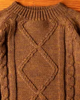 Buzo de lana merino color marrón con diseño de tejido trenzado en el frente y cuello redondo acanalado. Presenta puños y ruedo elásticos, con una pequeña etiqueta decorativa en la parte inferior.