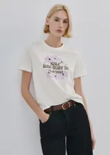 Remera de algodón color blanco, con cuello redondo y mangas cortas. Presenta un estampado frontal con flores en tonos lila y la frase "Self made ready to flourish" en letras negras.