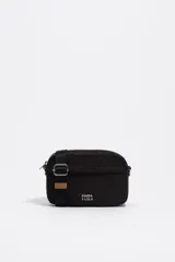 Bolso bandolera mini de nylon negro, con estructura blanda y cierre de cremallera. Posee una correa regulable de tejido negro con hebilla metálica plateada y un detalle de cuero sintético marrón en la correa. Lleva el logo metálico de Bimba y Lola en la parte frontal inferior.