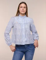 Blusa celeste de algodón con estampado floral, cuello mao fruncido con volado y cordel frontal con borlas. Tiene mangas largas abullonadas con puños elásticos y abotonadura frontal.
