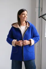 Tapado de paño color azul con capucha, cierre frontal y detalles en color beige en el interior de la capucha, puños y bordes de los bolsillos.