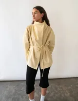 Campera de algodón color amarillo manteca, con cuello alto, cierre frontal con botones metálicos y cinturón ajustable en la cintura. Presenta un diseño versátil que permite llevarla entallada o con silueta holgada.