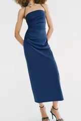 Vestido midi de color azul, con escote recto y cuello halter que incorpora una aplicación metálica tipo choker. Presenta un diseño con frunces laterales en el frente.