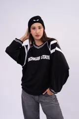 Buzo estilo jersey deportivo color negro, con cuello en V y detalles de franjas blancas en los hombros y puños. Presenta la inscripción 'Michigan State' estampada en el frente.