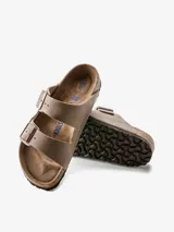 Sandalia tipo zueco Birkenstock modelo Arizona, color marrón topo, con dos tiras ajustables con hebillas metálicas color bronce. Presenta una plantilla anatómica de corcho y goma, y una capa adicional de espuma para mayor confort.