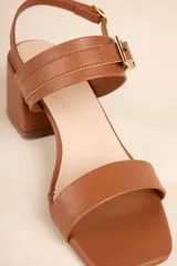 Sandalia negra de símil cuero con taco ancho y tiras ajustables con hebilla metálica. Presenta una tira ancha sobre los dedos y una tira fina con hebilla que sujeta el tobillo.