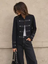 Chaqueta de estilo militar en color negro, confeccionada en tejido de lana. Presenta cuello redondo, cierre frontal con botones metálicos y detalles de tiras horizontales. Cuenta con mangas largas con puños decorados con botones y diseño estructurado.