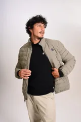 Campera puffer de corte clásico en color beige, con cierre frontal, cuello alto y puños elásticos. Presenta un diseño acolchado horizontal y un pequeño logo bordado en el pecho.