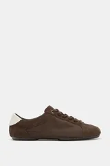 Zapatilla estilo blucher con diseño deportivo, confeccionada en una combinación de materiales textiles y gamuza en tono marrón, con detalle de contraste en color blanco en el talón y cierre mediante cordones.