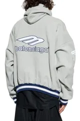 Hoodie Balenciaga de color gris claro con capucha, cierre frontal parcial con botones y cuello tipo polo. Presenta un escudo bordado en el pecho, estampa con la marca y gráficos deportivos en el frente y las mangas, además de puños y cintura elastizados con rayas azul y blanca.