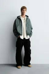 Cazadora negra de algodón con efecto lavado, corte relajado y capucha ajustable. Presenta cierre frontal de cremallera, bolsillos frontales tipo canguro y elásticos en puños y bajo.