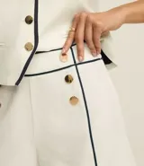 Short sastre de cintura alta confeccionado en crepé color blanco, con botones delanteros metalizados, cierre lateral por cremallera, bolsillos falsos atrás y bies contrastante en color negro.