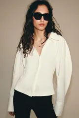 Top de tejido combinado color blanco, con cuello solapa, escote en V y cierre frontal con botones. Presenta mangas largas con puños anchos.