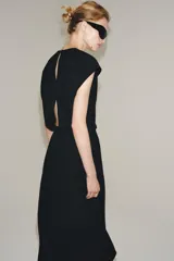 Vestido midi negro con cuello redondo y manga sisa, confeccionado por Zara Woman Collection. Presenta un diseño drapeado en la parte delantera y un bajo asimétrico con un voluminoso volante lateral. Incluye un pequeño cierre de botón en la espalda.