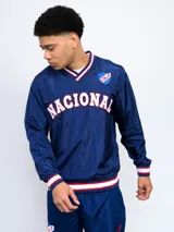 Buzo deportivo azul marino de nylon con cuello en V, puños y cintura acanalados con detalles tricolor (blanco, rojo y azul). Estampado frontal con la palabra "NACIONAL" en blanco y escudo del club C.N.de F. en el pecho. Corte clásico.