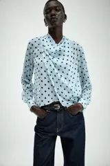 Camisa de manga larga con cuello fluido y diseño cruzado en el frente. Confeccionada en tejido semitransparente de color celeste con estampado de lunares negros. Presenta bajo asimétrico y cierre en la espalda con botones forrados.