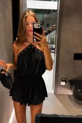 Vestido corto negro con diseño halter, cuello con ojales metálicos decorativos y cintura elástica fruncida. Presenta una falda con vuelo y caída fluida.
