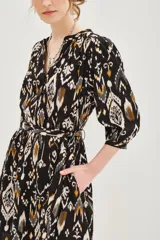 Vestido corto de corte recto con estampado ikat en tonos negro, blanco y ocre. Presenta escote en V con cierre de botones, mangas tres cuartos con puños elásticos y un cinturón de la misma tela para ajustar la cintura.
