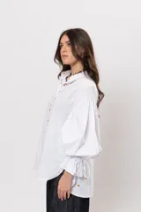 Camisa de algodón blanca de silueta amplia, con cuello camisero, botonadura frontal con botones decorativos, mangas abullonadas extragrandes y detalles de tiritas ajustables con cuentas de resina en los puños.