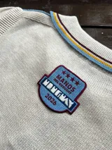 Remera de punto color crudo con cuello redondo y detalles de rayas en azul, amarillo y bordó. Presenta un parche bordado en el pecho con el logo de Manos del Uruguay y la inscripción 2026.