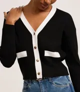 Cardigan de punto blanco con escote en V, botones metalizados, bolsillos falsos, cuello y vista contrastantes en negro, y bajo acanalado.
