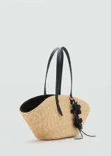 Bolso tipo shopper de fibra natural con diseño en forma de abanico. Cuenta con dos asas largas de hombro en color negro y un detalle decorativo de flores negras colgando de una de las asas.