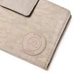 Billetera tipo fichero color beige, confeccionada en material textil con cierre de solapa y broche magnético. Incluye una correa de muñeca extraíble y un aplique circular con el logo de la marca en el frente.