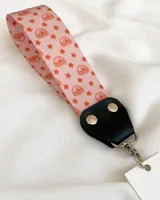 Llavero de muñeca con cinta de tela estampada con arcoíris y estrellas color naranja sobre fondo rosa. Tiene un conector de cuero sintético negro y un gancho metálico plateado.