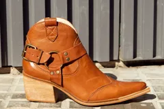 Bota tipo texana de caña baja confeccionada en cuero natural, con taco cuadrado de madera y punta afinada. Presenta una tira decorativa con hebilla metálica y estribo lateral, además de costuras reforzadas en el empeine.