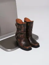 Botas estilo cowboy de cuero en tono marrón con efecto desgastado. Presentan un diseño de caña media con detalles de correas, argollas metálicas y tachas decorativas. Cuentan con un taco cuadrado de altura media y punta ligeramente cuadrada.