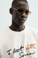 Camiseta blanca de algodón con corte boxy fit y cuello redondo. Presenta un estampado frontal con texto manuscrito en negro y marrón.