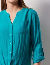 Blusa blanca de gasa marca Zac & Rachel, con cuello a la base, escote en V, abertura central con botones, bolsillo aplicado en el pecho y mangas largas regulables con presilla y botón. Tiene terminación opcional de nudo en el ruedo.