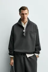 Sudadera de corte relajado color negro, con cuello alto y cierre frontal de cremallera. Presenta un bolsillo de vivo en el pecho con un estampado a contraste y acabados elásticos en puños y ruedo.