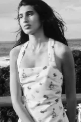 Vestido mini de color beige con un estampado de tomates, aceitunas y botellas de aceite, y un borde inferior rojo con ilustraciones de una sombrilla de playa y vegetales. Cuenta con escote cuadrado y tirantes anchos cruzados en la espalda.