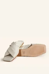 Sandalia tipo slide de cuero vacuno color blanco, con diseño de tiras cruzadas y fruncidas. Presenta una puntera cuadrada y una suela plana de cuero con un pequeño taco de 2 cm.