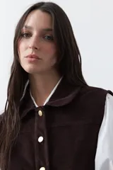 Chaleco de pana color marrón oscuro, con cuello camisero, cierre frontal de botones metálicos dorados y dos bolsillos frontales con detalle de bordado geométrico.