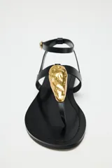 Sandalia plana negra con tira para el dedo y tira tobillera ajustable con hebilla dorada. Presenta un adorno grande de pieza metálica dorada con textura orgánica sobre el empeine.