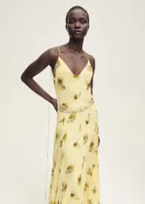 Vestido largo de tirantes finos con cuello de pico, confeccionado en viscosa con estampado floral difuminado en tonos amarillos y marrones. Presenta un diseño recto con detalle de cadena metálica a la altura de la cadera y cierre de lazo en la espalda.