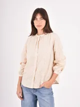 Camisa de manga larga en color crudo, confeccionada en tela con delicado bordado floral en relieve. Presenta cuello redondo con frunces, cierre frontal con botones y mangas abullonadas con puños ajustados.