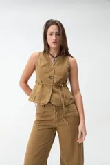 Chaleco de denim color beige, sin mangas, con escote en V, cierre frontal con botones y lazo ajustable en la cintura.