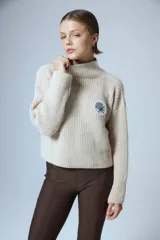 Sweater de punto grueso color crema, con cuello alto y parche circular con bordado en el pecho.
