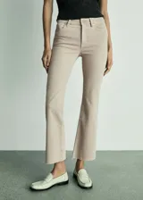 Jeans flare tiro alto color beige, de diseño crop con bajo deshilachado, cinco bolsillos, trabillas para cinto y cierre frontal con cremallera y botón.