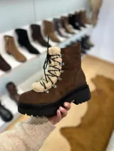Bota tipo borcego de cuero marrón con suela track negra de goma. Presenta un aplique frontal desmontable de corderito sintético color crema y cordones negros con ganchos metálicos.
