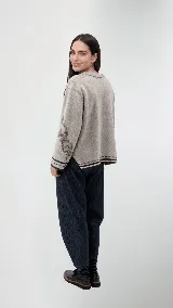 Sweater de lana color beige con calce holgado y cuello redondo. Presenta un bordado negro con diseño de arabescos en una de las mangas y detalles de rayas y puntos en contraste en el ruedo y los puños.