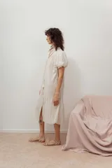 Vestido midi color crudo, con cierre frontal de botones, escote en V y mangas cortas abullonadas. Presenta un diseño holgado con detalle de volados en el bajo.